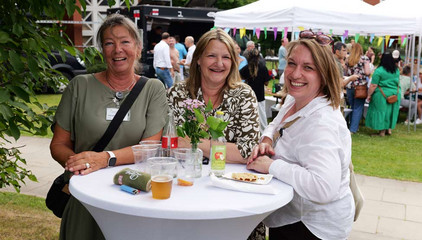Drei Mitarbeiterinnen stehen an einem mit weißem Stoff bezogenen Stehtisch mit Blumenschmuck und Getränken und lächeln in die Kamera beim Sommerfest 2026 auf dem Gesundheitscampus Volksdorf des Evangelischen Amalie Sieveking Krankenhauses, der Residenz am Wiesenkamp, des Feierabendhauses Volksdorf und der Albertinen Kindertagesstätte Volksdorf. 