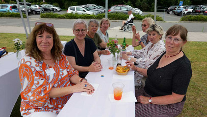 Sieben Besucherinnen sitzen an einem mit weißem Stoff überzogenen Tisch mit Getränken, Essen und Blumenschmuck und blicken in die Kamera beim Sommerfest 2026 auf dem Gesundheitscampus Volksdorf des Evangelischen Amalie Sieveking Krankenhauses, der Residenz am Wiesenkamp, des Feierabendhauses Volksdorf und der Albertinen Kindertagesstätte Volksdorf. 