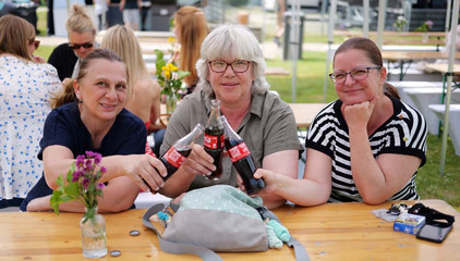 Drei Mitarbeiterinnen sitzen an einem Tisch und stoßen mit Colaflaschen an beim Sommerfest 2026 auf dem Gesundheitscampus Volksdorf des Evangelischen Amalie Sieveking Krankenhauses, der Residenz am Wiesenkamp, des Feierabendhauses Volksdorf und der Albertinen Kindertagesstätte Volksdorf. 