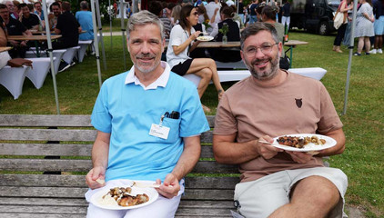 Zwei Mitarbeiter sitzen mit vollen Tellern auf einer Sitzank und blicken in die Kamera beim Sommerfest 2026 auf dem Gesundheitscampus Volksdorf vom Evangelischen Amalie Sieveking Krankenhaus, Residenz am Wiesenkamp, Feierabendhaus Volksdorf und Albertinen Kindertagesstätte Volksdorf.