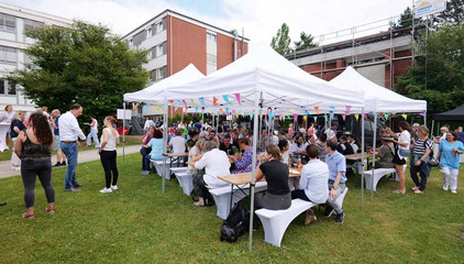 Besucherinnen und Besucher sitzen unter weißen Pavillons an Tischen und Sitzbänken auf einer Rasenfläche beim Sommerfest 2026 auf dem Gesundheitscampus Volksdorf des Evangelischen Amalie Sieveking Krankenhauses, der Residenz am Wiesenkamp, des Feierabendhauses Volksdorf und der Albertinen Kindertagesstätte Volksdorf.