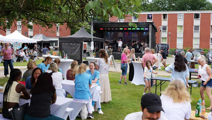 Menschen sitzen draußen auf dem Außengelände mit Tischen, Sitzbänken, Zelten und Musikbühne beim Sommerfest 2026 auf dem Gesundheitscampus Volksdorf vom Evangelischen Amalie Sieveking Krankenhaus, Residenz am Wiesenkamp, Feierabendhaus Volksdorf und Albertinen Kindertagesstätte Volksdorf. 